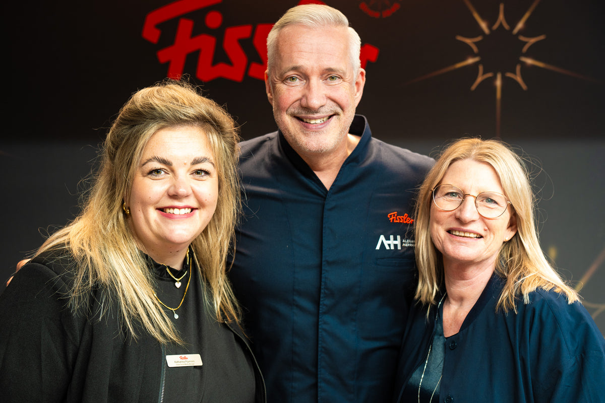 Three adults, two women and a chef, smile in the kitchen. The Fissler logo and starry sky emphasize the tradition of our premium cookware made in Germany.