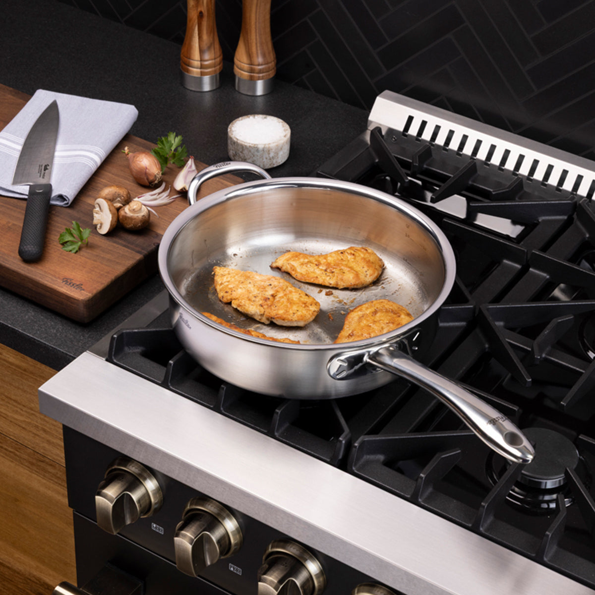 Drei Hähnchenbruststücke brutzeln in einer Fissler M5 Pro-Ply® Sautépfanne mit Metalldeckel auf einem Gasherd und verdeutlichen damit die präzise Technik und Langlebigkeit unseres in Deutschland hergestellten Edelstahl-Kochgeschirrs. Neben dem Herd liegt auf einem Schneidebrett ein hochwertiges Fissler-Messer neben frischen Pilzen, Petersilie und einem ordentlich gefalteten Handtuch. Die Szene spielt sich in einer modernen Küche mit einer eleganten schwarzen Küchenrückwand ab, die Fisslers Engagement für