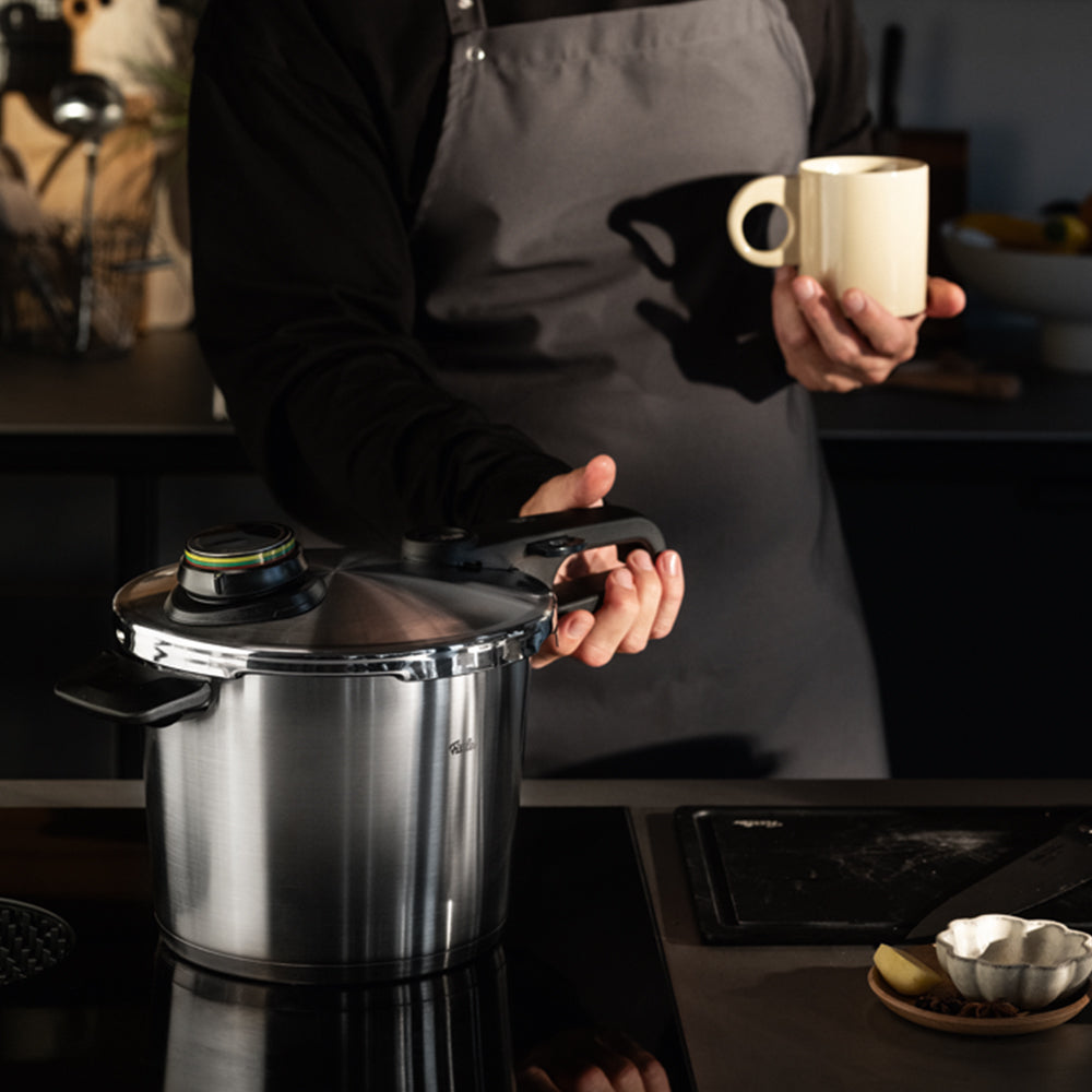 Eine Person in einer grauen Schürze steht in einer modernen Küche, hält einen gelben Becher in der Hand und präsentiert einen hochwertigen Fissler-Edelstahl-Schnellkochtopf. Im Hintergrund sind verschiedene Küchenutensilien und Geschirr zu sehen, die Fisslers Engagement für Präzisionstechnik, Langlebigkeit und zeitloses deutsches Design widerspiegeln.