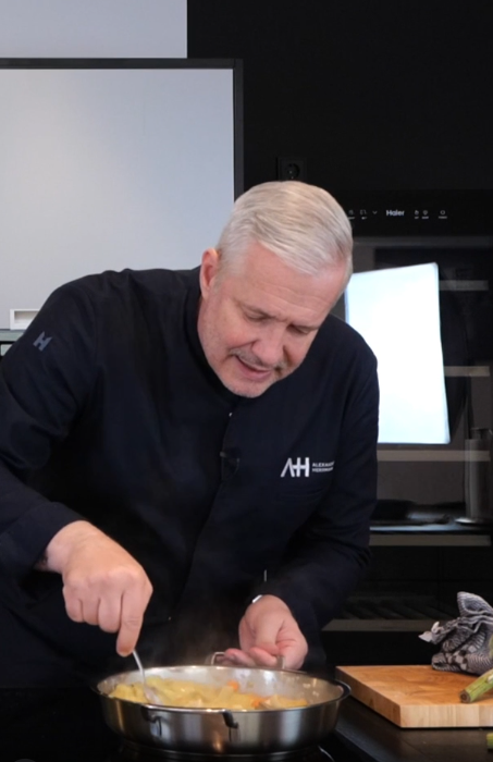 Ein Mann in einer dunklen Kochjacke bereitet ein Gericht mit Fissler-Edelstahlkochgeschirr auf einem Herd in einer eleganten modernen Küche zu.