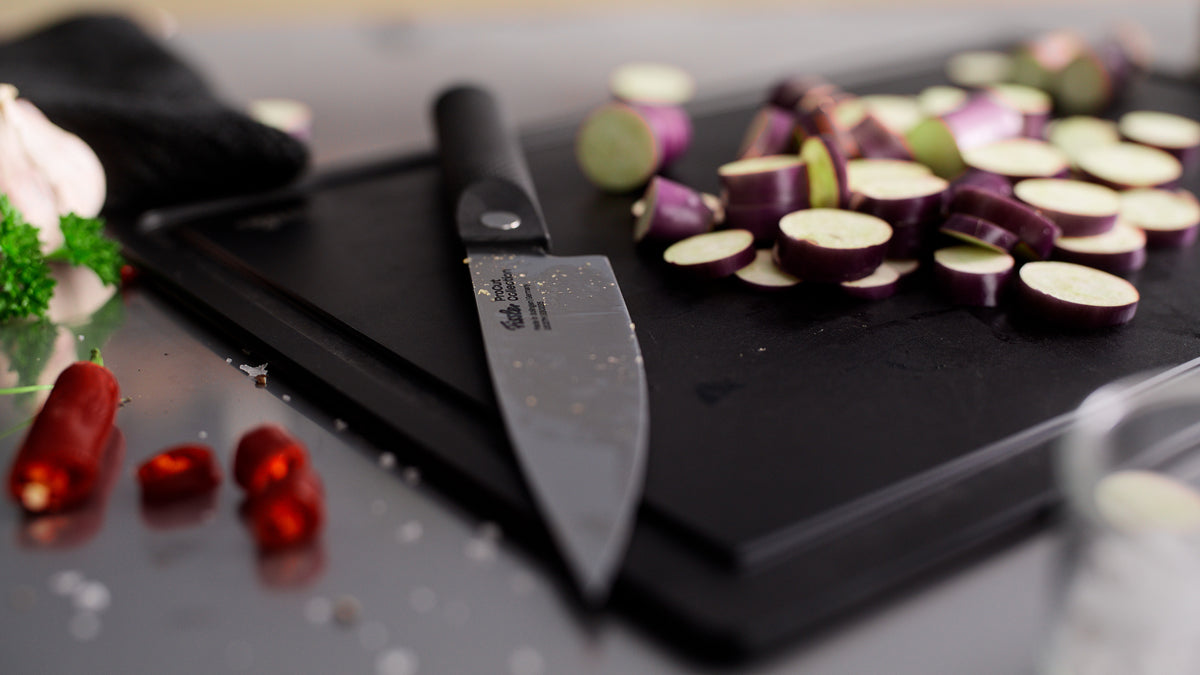 Das Fissler ProCut Kochmesser (16 cm) besticht durch präzise Handwerkskunst und einen eleganten schwarzen Griff und ist aus extrahartem Stahl gefertigt, der für lange Haltbarkeit sorgt. Das Messer wird auf einem schwarzen Schneidebrett neben frisch geschnittenen violetten Auberginenscheiben präsentiert, zusammen mit leuchtend roten Chilischoten und frischen Kräutern auf der Küchenarbeitsplatte – ein Beweis für Fisslers Tradition hochwertiger deutscher Kochutensilien seit 1845.