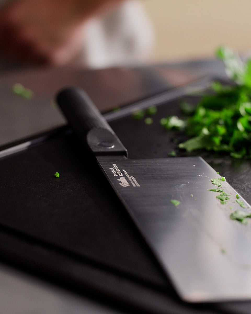 Eine Nahaufnahme des Fissler ProCut Chinesisches Kochmessers (18 cm) mit robustem schwarzem Griff und einer Klinge aus extrahartem Stahl liegt auf einem Schneidebrett neben frisch gehackten grünen Kräutern – ideal zum präzisen Schneiden von Kräutern und Gemüse. Im Hintergrund ist eine verschwommene Hand zu sehen. Als Beispiel für Fisslers Tradition der Präzisionstechnik und des zeitlosen deutschen Designs seit 1845 unterstreicht dieses Messer unser Engagement für langlebige, hochwertige Küchenutensilien.