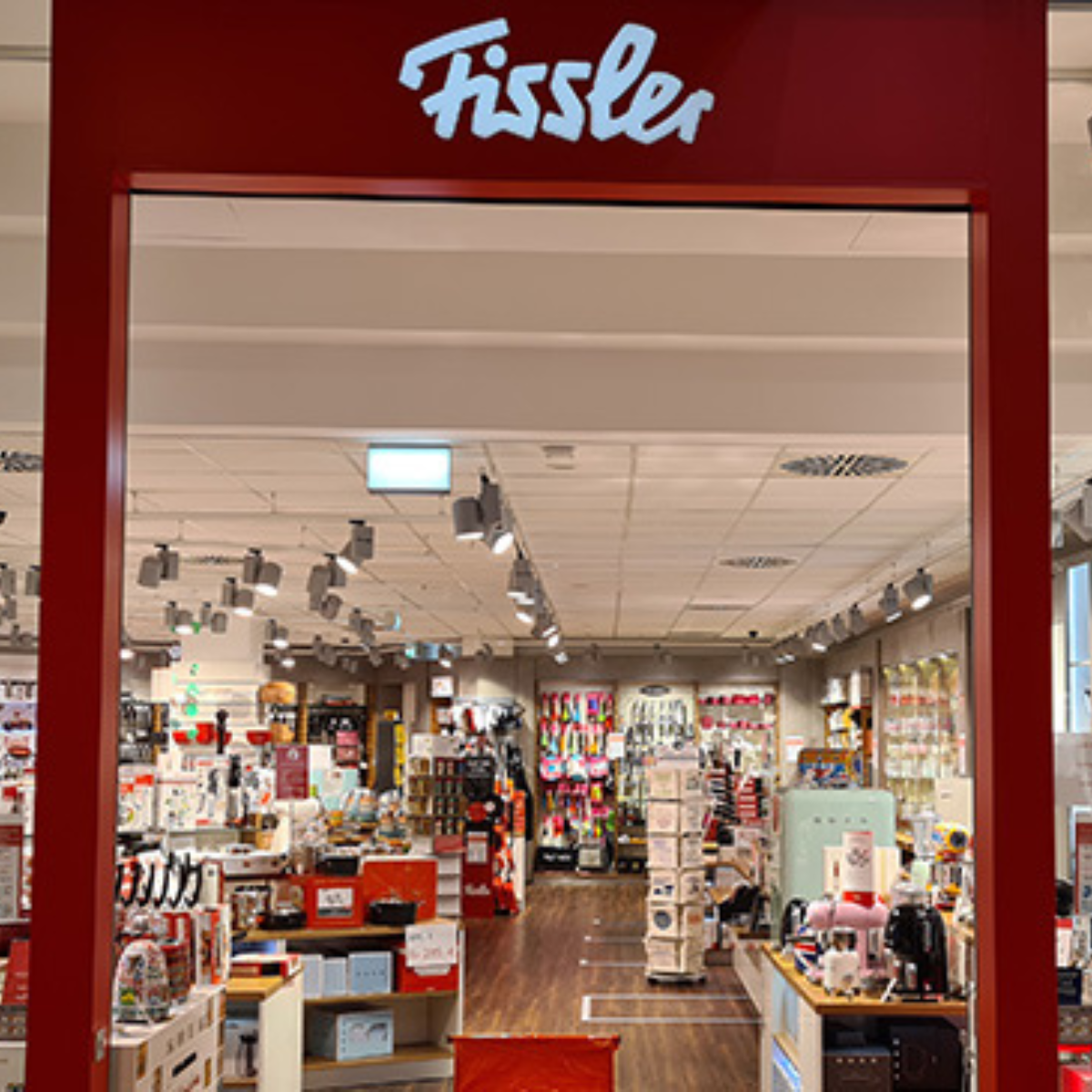 Das Innere eines Fissler-Geschäfts präsentiert Kochgeschirr und Zubehör aus rostfreiem Stahl aus deutscher Produktion durch einen rot umrahmten Eingang, Logo oben.