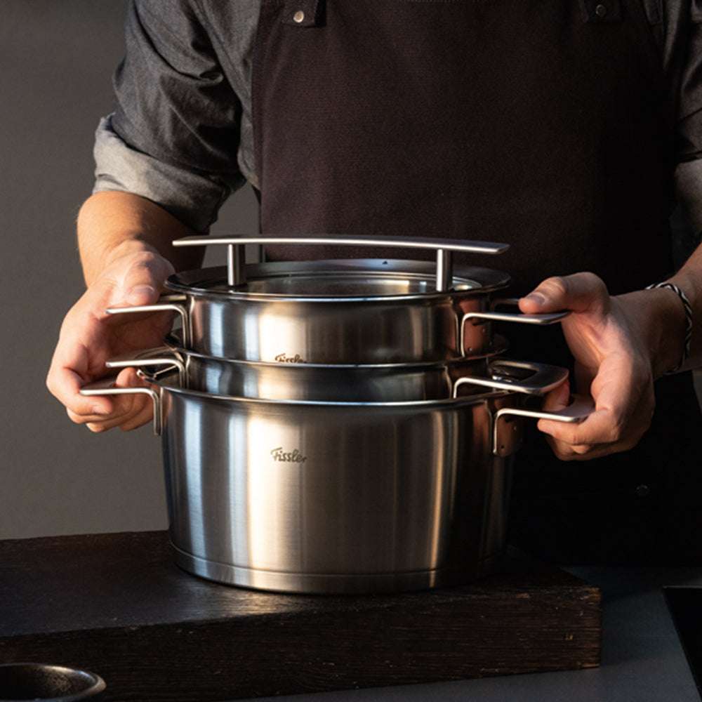Eine Person in einer dunklen Schürze stapelt zwei Fissler-Edelstahltöpfe mit ergonomischen Griffen auf einer eleganten, dunklen Oberfläche und unterstreicht damit die bekannte deutsche Präzision und das zeitlose Design der Marke.