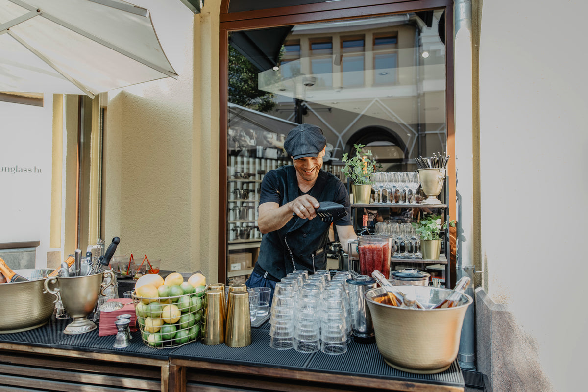 Ein Barkeeper in schwarzer Uniform und mit schwarzer Mütze bereitet an einer Außenbar gekonnt Getränke zu, umgeben von präzise gefertigten Fissler-Barartikeln aus Edelstahl, frischem Obst, Gläsern und Flaschen. Das elegante Ambiente spiegelt zeitloses deutsches Design und Innovation wider, während im Hintergrund ein Gebäude elegant zu sehen ist.