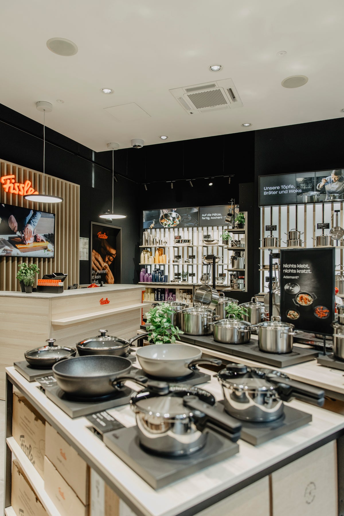 Der Fissler-Showroom besticht durch seine Innenausstattung mit präzise gefertigten Edelstahltöpfen, -pfannen, -Schnellkochtöpfen und Küchenzubehör, die elegant auf Arbeitsplatten und Regalen angeordnet sind. Warme Beleuchtung unterstreicht die Langlebigkeit und das zeitlose Design des Kochgeschirrs „Made in Germany“ und schafft so ein einladendes und stilvolles Einkaufserlebnis.