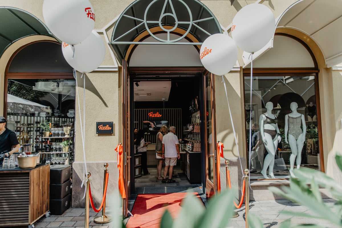 Eingang zum Geschäft mit Fissler-Schild, rotem Teppich und weißen Luftballons. Zwei Personen betreten das Geschäft. Im Schaufenster sind elegante Auslagen mit hochwertigem Kochgeschirr aus Edelstahl zu sehen. Eine Auslage im Außenbereich auf der linken Seite präsentiert präzise gefertigte Töpfe und Pfannen, die Fisslers Tradition von Qualität und zeitlosem Design widerspiegeln.