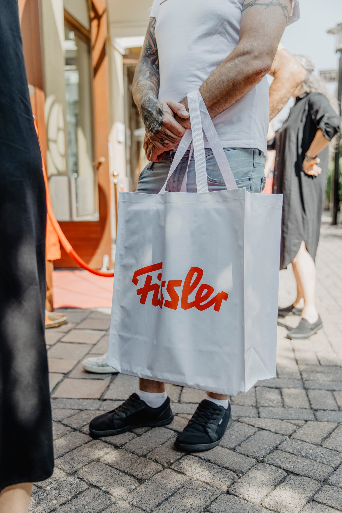 Eine Person mit tätowierten Armen, gekleidet in ein weißes T-Shirt und Jeans, steht auf einem gepflasterten Gehweg und hält eine weiße Tragetasche mit dem ikonischen roten Fissler-Logo in der Hand – ein Symbol für deutsche Präzisionstechnik und zeitloses Design. Im Hintergrund sind andere Personen unscharf zu sehen, wodurch die Fissler-Tasche besonders hervorgehoben wird, die für unsere Tradition hochwertiger Kochgeschirre aus Edelstahl und innovativer Küchenaccessoires steht.
