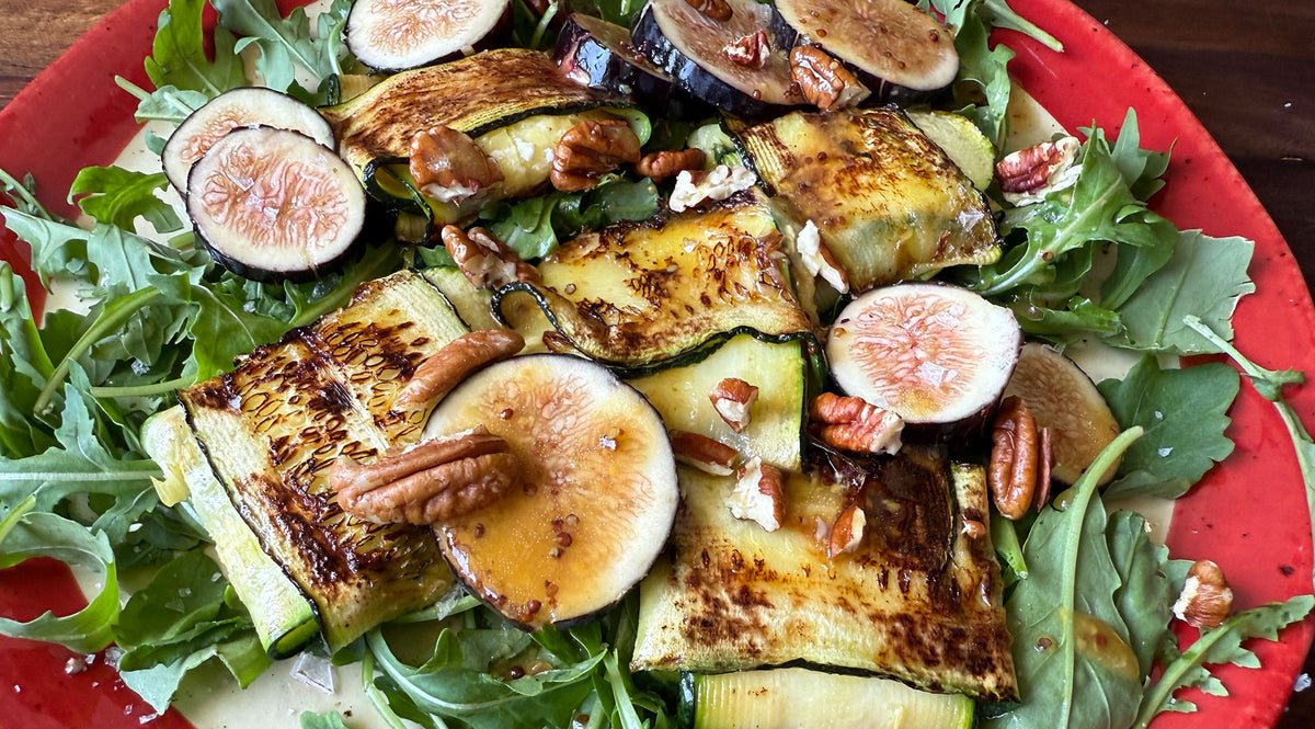 Ein farbenfroher Salat aus gegrillten Zucchinischeiben, frischem Rucola, in Scheiben geschnittenen Feigen und Pekannüssen, elegant auf einem roten Teller angerichtet und mit einem Spritzer Balsamico-Vinaigrette verfeinert – präsentiert auf dem präzise gefertigten Kochgeschirr von Fissler, das zeitlose deutsche Handwerkskunst widerspiegelt.