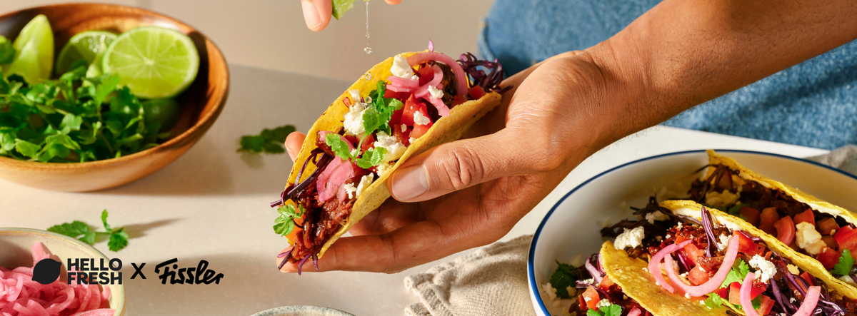 A hand squeezes fresh lime over vibrant tacos filled with vegetables and cheese, with extra lime wedges and cilantro arranged nearby. The scene is set on premium Fissler stainless steel cookware, reflecting German precision engineering and timeless design. “Hello Fresh x Fissler” logos appear in the bottom left corner, highlighting quality kitchen performance.