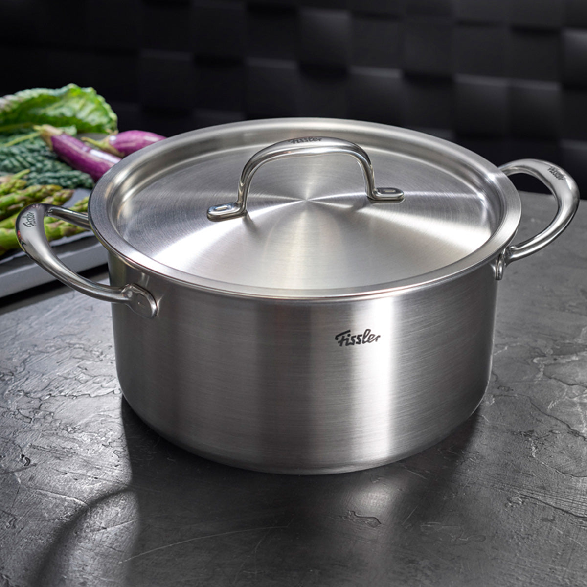 Das 4-teilige Fissler M5 Pro-Ply®-Set mit Metalldeckeln aus recyceltem Edelstahl „Made in Germany“ präsentiert sich auf einer dunklen Arbeitsplatte. Umgeben von stilvoll arrangiertem grünem und violettem Gemüse unterstreicht das innovative, präzise verarbeitete Kochgeschirr unsere Tradition für langlebige Qualität und zeitloses Design. Das vielseitige Set ist die ideale Ergänzung zu jedem Pfannenset und eignet sich optimal für alle induktionsgeeigneten Küchen.