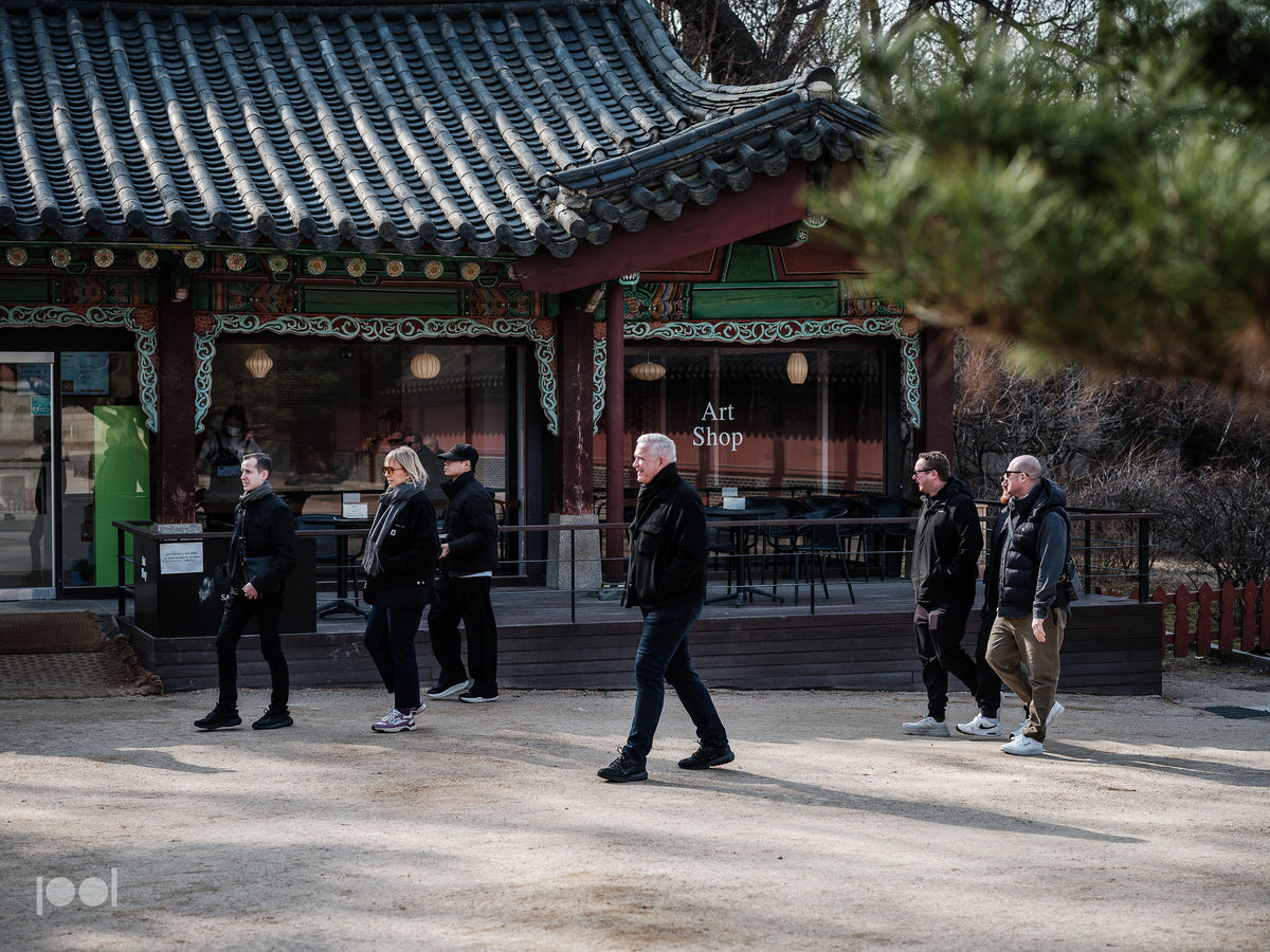 Six people stroll past a traditional Korean building with ornate roof tiles, evoking timeless craftsmanship. The structure, featuring an Art Shop sign, large glass windows, and tables and chairs inside, stands amid lush greenery. The scene reflects Fissler’s dedication to enduring design and precision—qualities found in every piece of our German-made cookware: stainless steel pots, pans, pressure cookers, and kitchen accessories that honor both heritage and innovation.