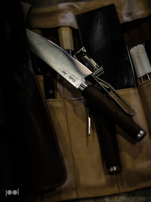A Fissler chef’s knife with a dark, ergonomically designed handle lies on a tan knife roll, accompanied by high-quality kitchen tools—a precision peeler, a reliable sharpening rod, and a robust black cleaver—exemplifying Fissler’s commitment to German craftsmanship and timeless design.