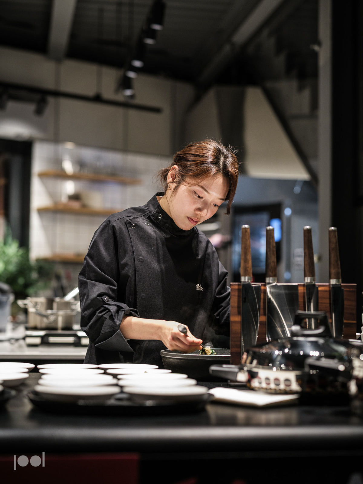 A chef in a black uniform prepares food with precision in a modern kitchen, focusing intently. Stainless steel Fissler pots, pans, and expertly crafted knives are arranged on the counter alongside other high-quality Fissler kitchen tools, reflecting German engineering and timeless design.